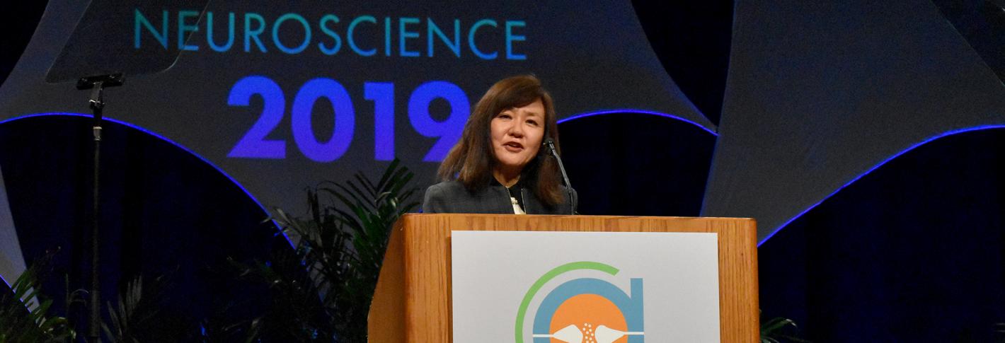 Li-Huei Tsai speaks from the podium with a huge Neuroscience 2019 sign behind her