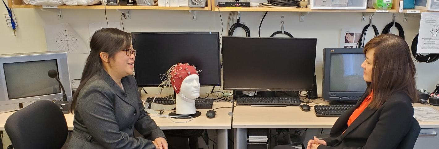 Diane Chan and Li-Huei Tsai sit in chairs facing eachother as they talk in a lab full of computer monitors and other electronics including a styrofoam head with an EEG cap on.