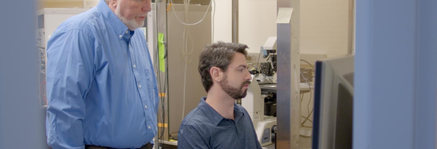 Andres Crane sits at a lab computer while Troy Littleton stands behind him looking over his shoulder at the screen.
