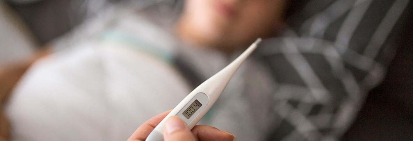 A thermometer shows a fever-level reading in the foreground as a sick child lies in the background