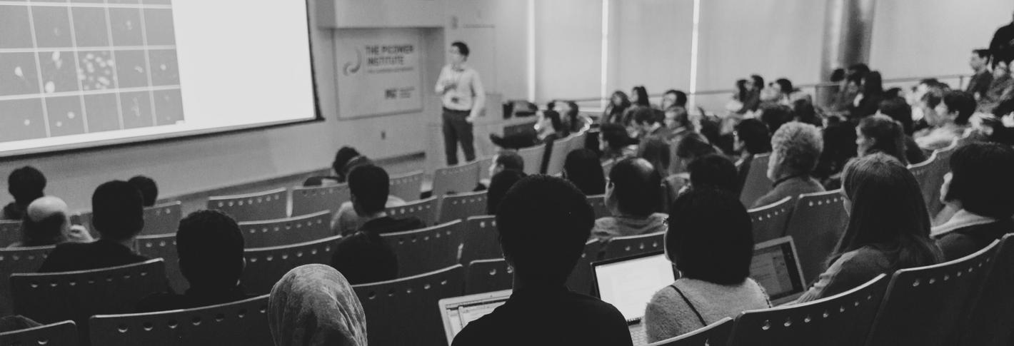 A black and white shot of a speaker and audience shot from the back of Singleton Auditorim