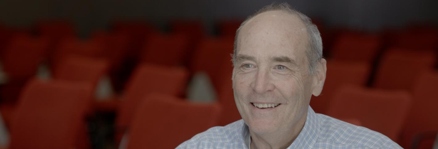 Mark Bear, shown from mid-chest up, smiles as he sits in a lecture room full of red chairs in the backgorund