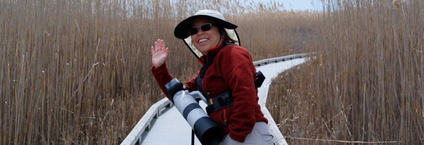 Ruby Lam waves to the camera as she stands on a boardwalk through tall grass and holds a camera with a large telephoto lens