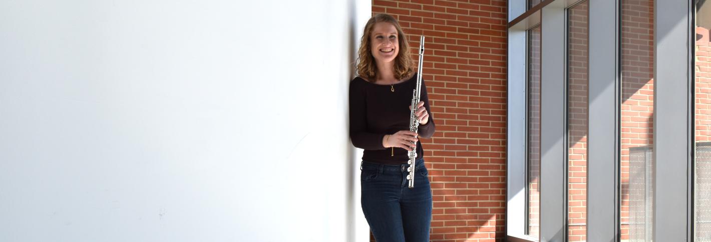 Sara Kornfeld-Sylla holds her flute as she stands along a curved white wall opposite floor to ceiling windows in a bright theater lobby