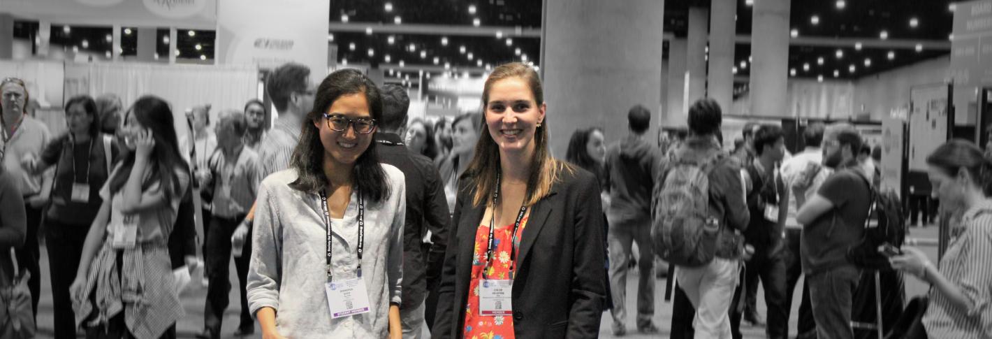 Two picower researchers are highlighted in color before a backdrop of the conference show floor, which is blurred and grayscale