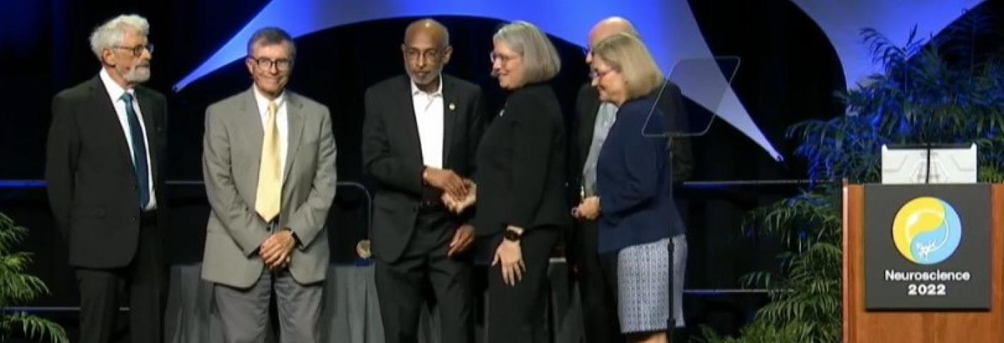 On a stage with plants and a blue background with a podium labeled Neuroscience 2022, Emery Brown receives a handshake from a woman after accepting a neuroscience prize