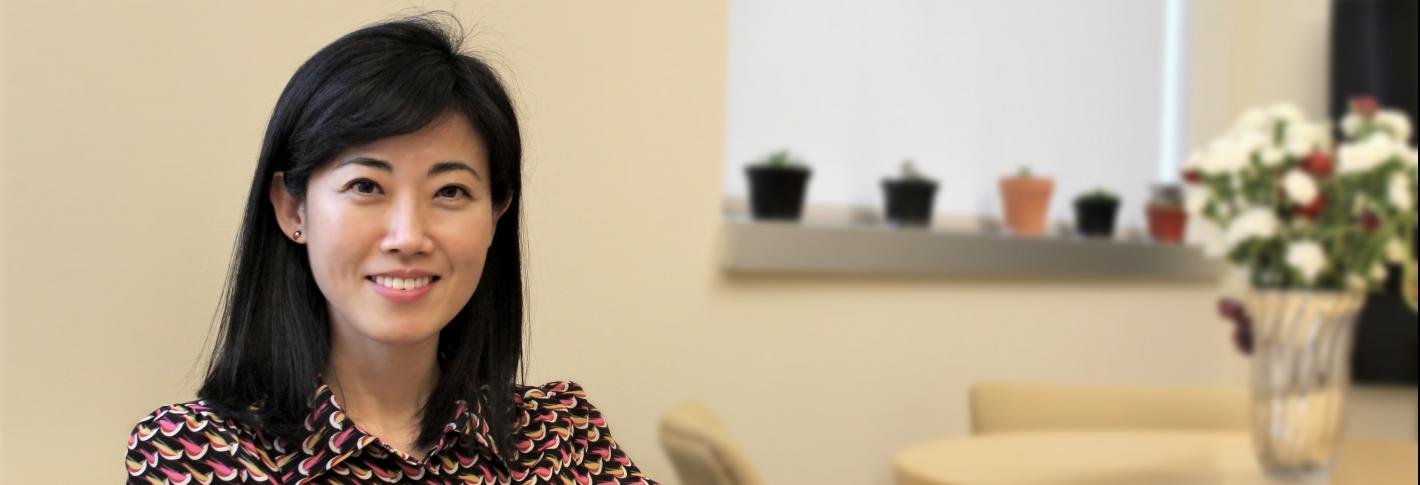 Gloria Choi smiles as she sits on a sofa in her office. The background. with flowers on a table. is blurred.