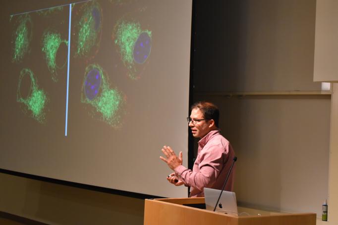 Eduardo Torres gestures as he stands between the podium and the screen which displays blue and green stained cell images