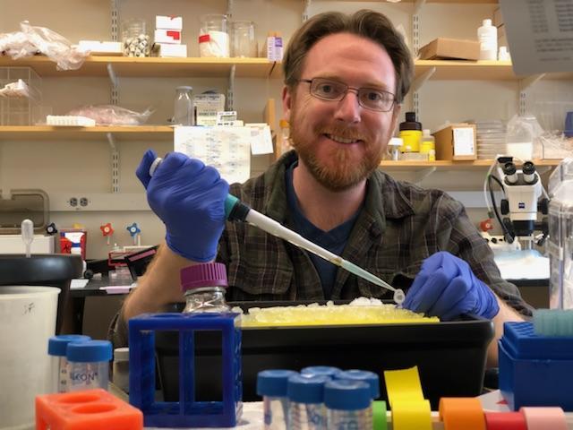 Patrick McCamphill smiles at hte camera as he holds a pipette in the lab