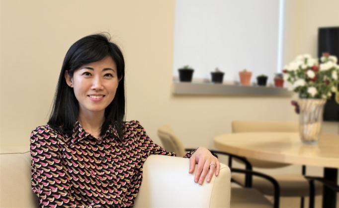 Gloria Choi smiles as she sits on a sofa in her office. The background. with flowers on a table. is blurred.