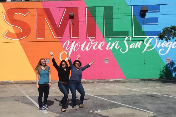 Three Picower researchers stand with arms happily in the air in a parking lot before a mural reading "Smile You're in San Diego"