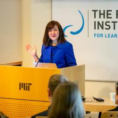 Li-Huei Tsai stands at a podium embossed with an MIT logo. In the foreground is an attentive audience. In the background is the Picower Institute logo