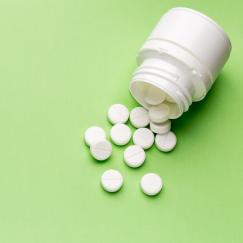 On a green background a white bottle of pills is overturned. Small round white pills have spilled out.