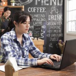 A young woman sits at a wooden table at a cafe staring at and typing on her laptop. The cafe counter is behind her and lots of menu options are written in chalk on a black menu board.