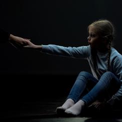 A little girl wearing blue sits on the floor in the dark. She reaches out to take an outstretched hand.