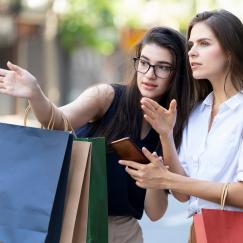 Two women on the street look intently and point in a particular direction. One holds a phone. The other holds shopping bags.