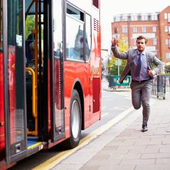 A man in a suit on a city street runs to catch up to a red bus at a stop.