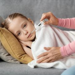 A little girl lies on a gray sofa under a blanket. A woman holds her gently with one hand and holds a thermometer in the girl's mouth with the other.