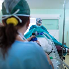An operating room scene shows a patient on a table. Our perspective is from behind the anesthesiologist who holds a mask on the patient's face and watches a monitor with a bunch of indicators. A surgeon stands out of focus on the far end of the patient.