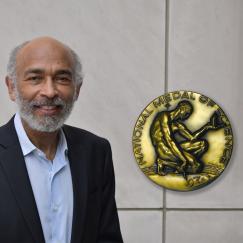 Emery N. Brown stands in front of a wall. Superimposed on the image next to him is a picture of the front of the National Medal of Science, which depicts a man kneeling and drawing an abstract character in the ground next to him as he contemplates an object.