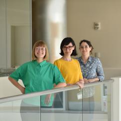 Taylor Baum, Josefina Correa Menendez and Karla Alejandra Montejo stand in a line and smile along a glass railing in a corridor of MIT Building 46.