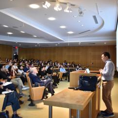 Li Ye is seen from the side as he delivers his talk at a symposium in MIT's Singleton Auditorium. In front of him is a nearly full room of audience members listening attentively.