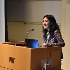 Gloria Choi stands at a wooden lectern with an MIT sign on it.
