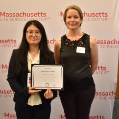 Shanshan Zhang and Rosie Firenze stand before a Massachusetts Life Sciences Center backdrop holding and award certificate