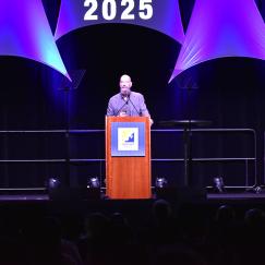 Earl Miller stands at the podium decorated with the Neuroscience 2025 logo and with audience members sitting in the foreground.