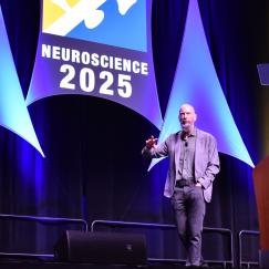 Earl Miller gestures walks across a large stage decorated with the words "Neuroscience 2025"