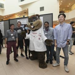 In the spacious, white atrium of MIT Building 46 several young men smile as they stand around MIT's Tim the Beaver mascot. Tim wears a white lab coat and has his hands out at his sides in a joyous gesture.