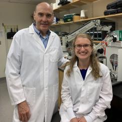 Wearing white lab coats in the lab, Mark Bear stands behind Sara Kornfeld-Sylla as she sits in a stool at a lab bench