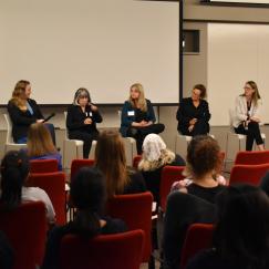 Six women, many holding microphones, sit in a row facing an audience of people sitting in red chairs.