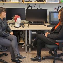 Diane Chan and Li-Huei Tsai sit in chairs facing eachother as they talk in a lab full of computer monitors and other electronics including a styrofoam head with an EEG cap on.
