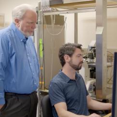 Andres Crane sits at a lab computer while Troy Littleton stands behind him looking over his shoulder at the screen.