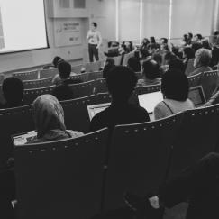 A black and white shot of a speaker and audience shot from the back of Singleton Auditorim