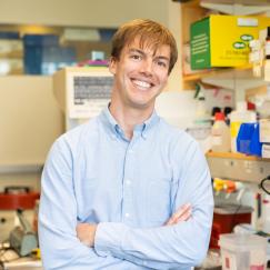 Smiling, arms crossed and wearing a light blue shirt, Mitch Murdock stands in front of a lab bench full of equipment