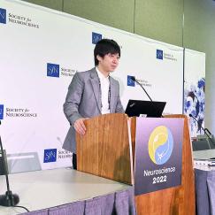 Takato Honda stands at a podium labeled Neuroscience 2022. Behind him is a white wall decorated with Society for Neuroscience logos