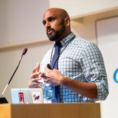 Ravi Raju stands at a podium. A blue and white Picower Institute logo is behind him as a backdrop.