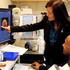 Li-Huei Tsai stands in a lab and points to an image of a cell on a computer screen. Hiruy Meharena sits at the desk with his hand on the mouse and looks on.