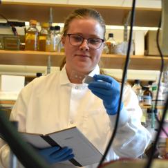 Eli Hanechak stands at a lab bench holding a notebook and looking at a slide. She is wearing a lab coat and blue latex gloves