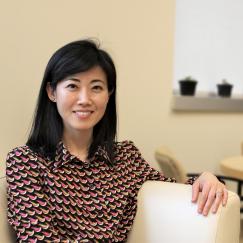 Gloria Choi smiles as she sits on a sofa in her office. The background. with flowers on a table. is blurred.
